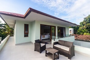 A modern house with a balcony, furnished with wicker furniture, and a bedroom visible through a glass door.