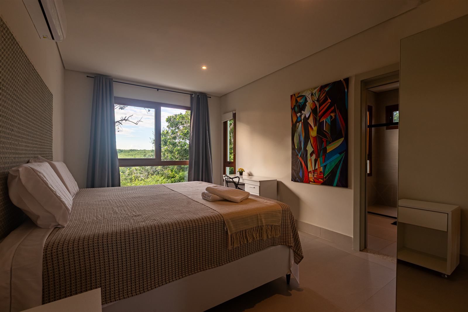 Een moderne slaapkamer met een groot bed, uitzicht op groen, een schilderij en een badkamer met douche.