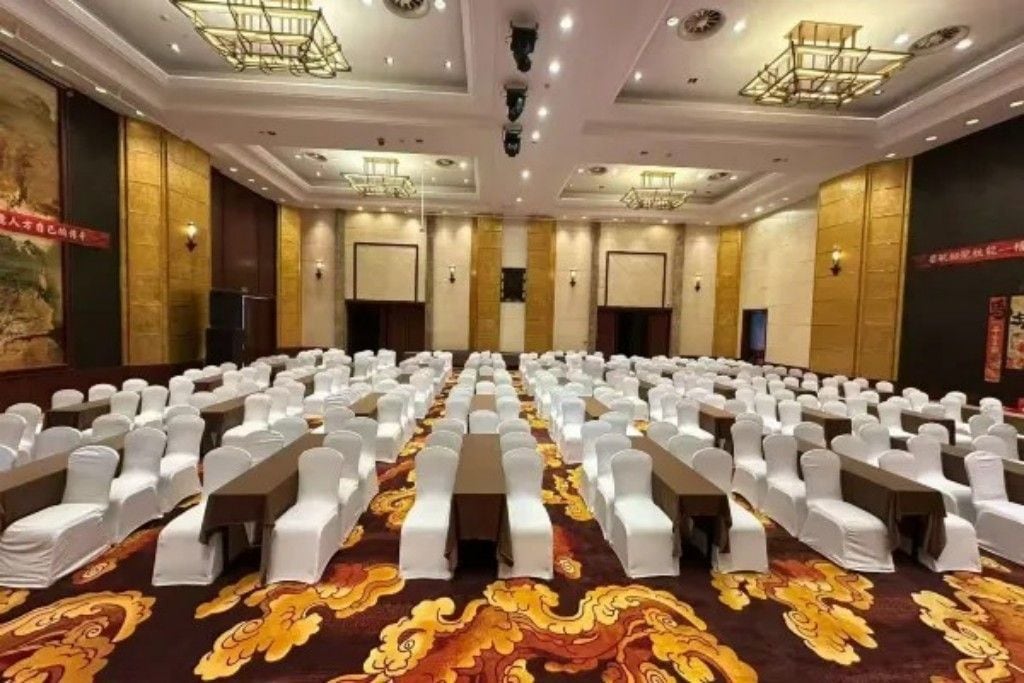 A conference room with rows of tables and chairs, a patterned carpet, and a ceiling with recessed lighting.