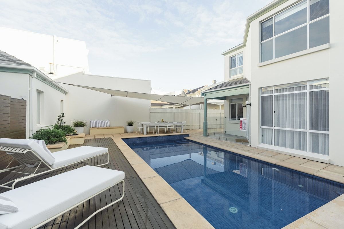 A swimming pool with lounge chairs, a table, and a covered seating area.
