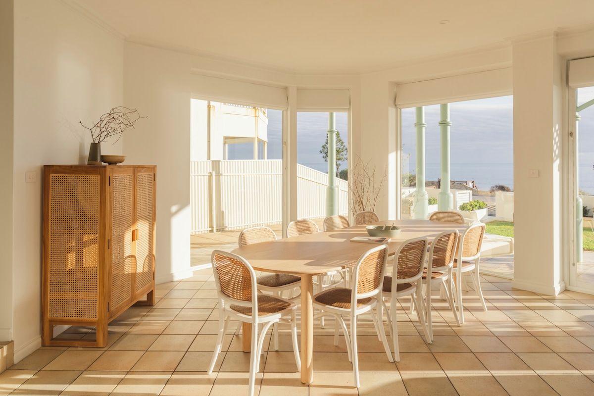Spiseområde med bord og stole, et fletskab og store vinduer med udsigt.