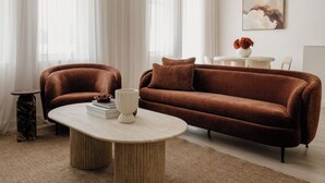 Un salon moderne avec un canapé marron, une table basse ronde et une grande fenêtre ornée de rideaux légers.
