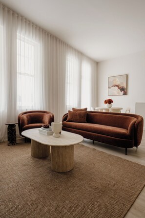 A modern living room with a brown sofa, a round coffee table, and a large window with sheer curtains. - Ilios | Luxury Apartment Steps from Henley Square (Henley Beach)
