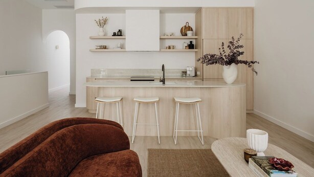 Une cuisine moderne avec un îlot central, des tabourets de bar blancs et un canapé marron, le tout dans un intérieur lumineux et minimaliste.