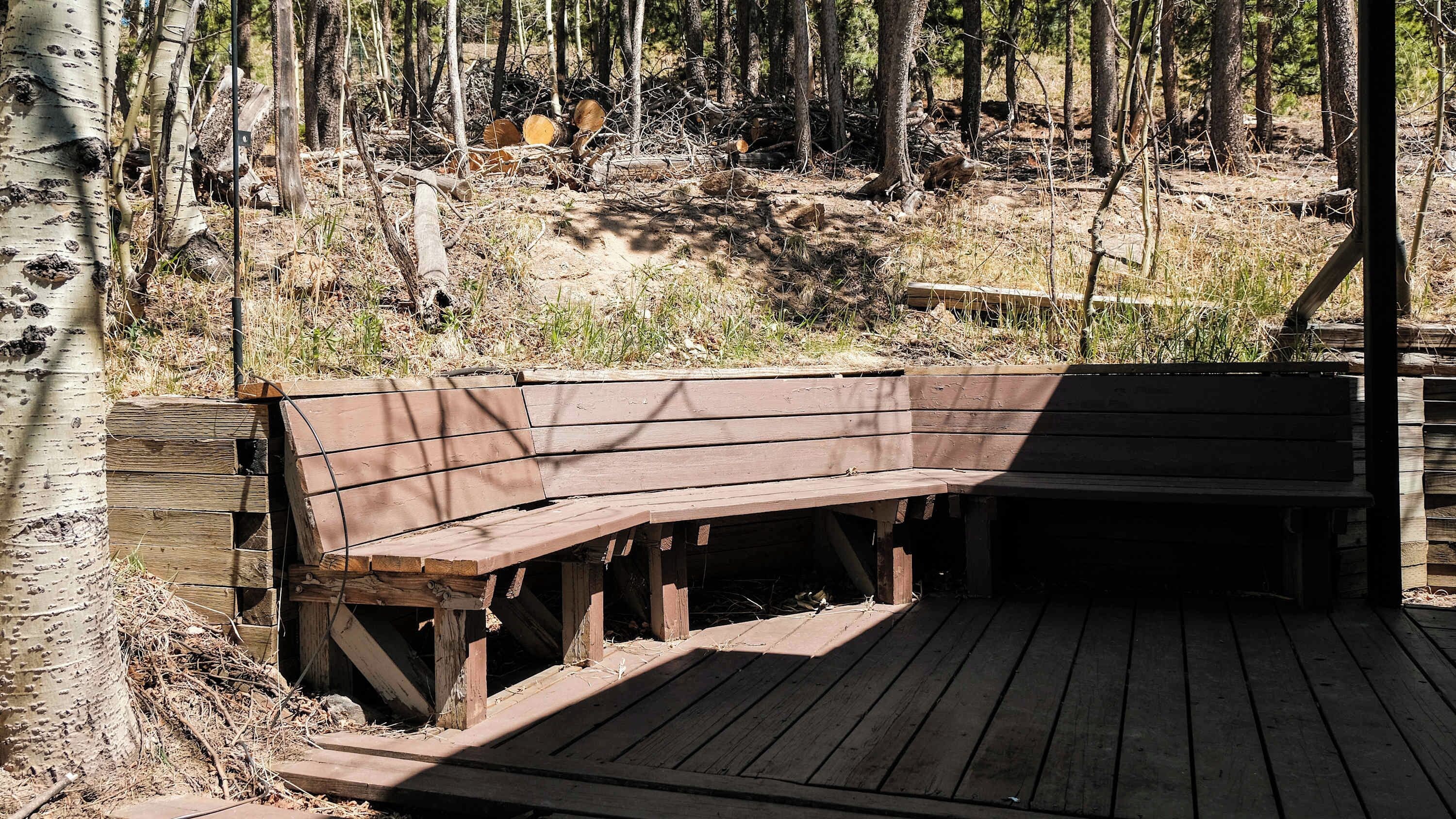 Un banco de madera con respaldo y reposabrazos, ubicado en una terraza de madera con un bosque natural de fondo.
