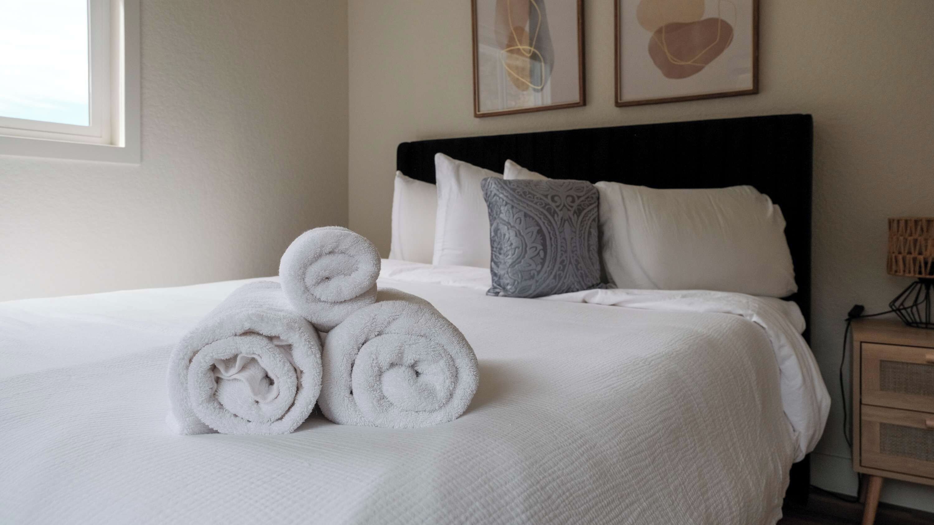 Un lit bien fait, avec des serviettes blanches pliées en rouleau, un coussin à motifs et une table de chevet surmontée d’une lampe.