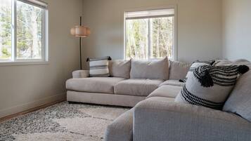 Un salon avec un canapé gris, un coussin décoratif à motifs, une lampe sur pied et une grande fenêtre donnant sur des arbres.