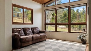 A room with a brown leather sofa, large windows with a view of trees, and a potted plant. - Hilltop Haus: Sweeping Mountain Views Above Denver (Morrison)