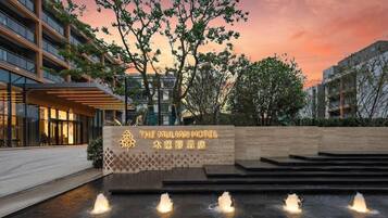 The exterior of The Mulian Hotel with a sign, a water feature, and a sunset in the background.