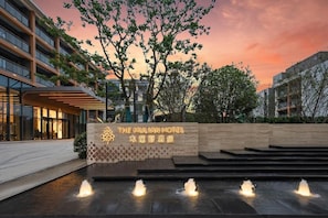 The exterior of The Mulian Hotel with a sign, a water feature, and a sunset in the background.
