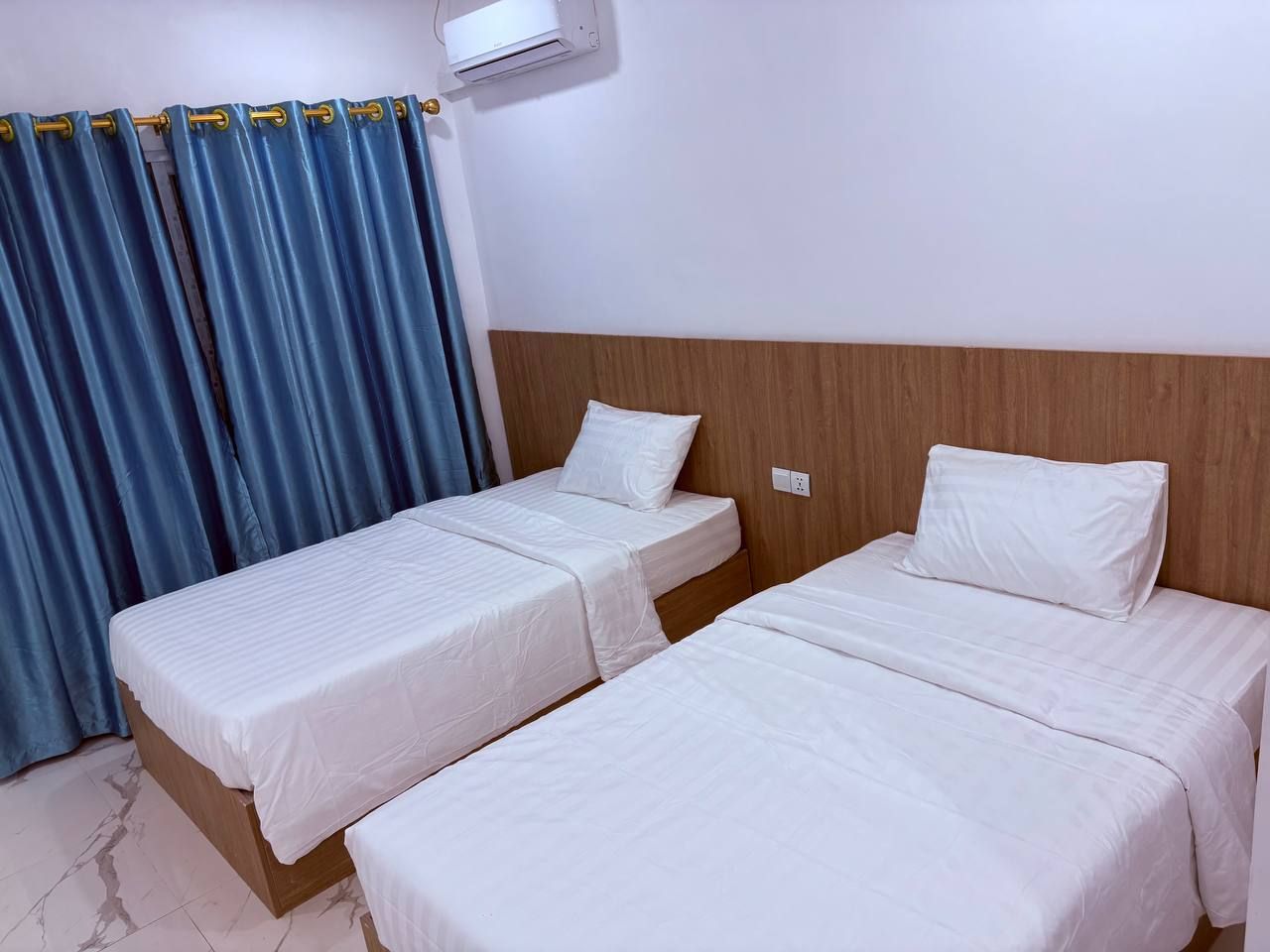 Une chambre d’hôtel avec deux lits simples, une tête de lit en bois, une fenêtre avec des rideaux bleus et un climatiseur fixé au mur.