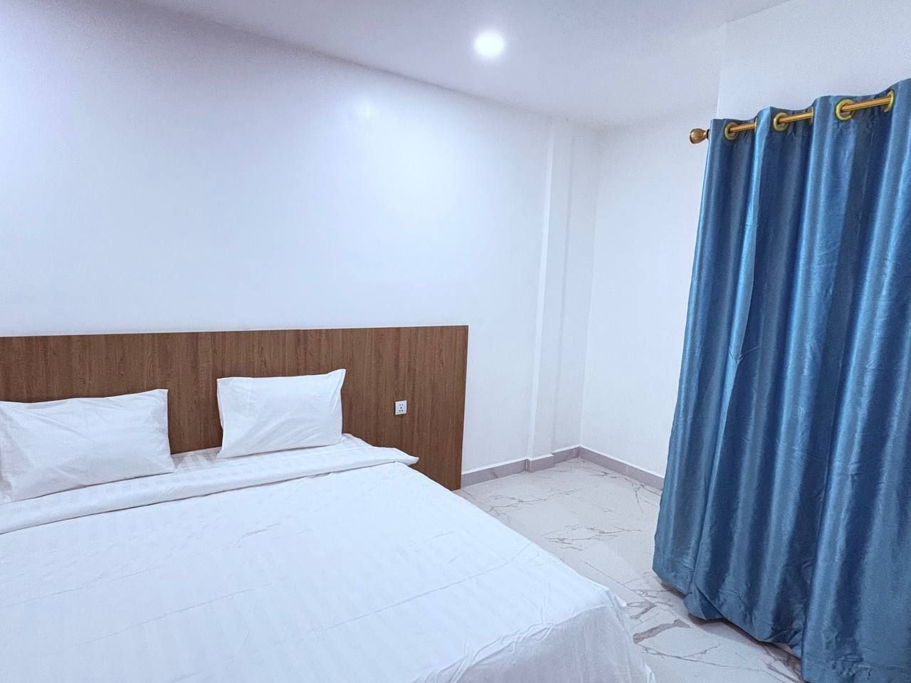 A hotel room with a bed, white bedding, a wooden headboard, a blue curtain, and a marble floor.