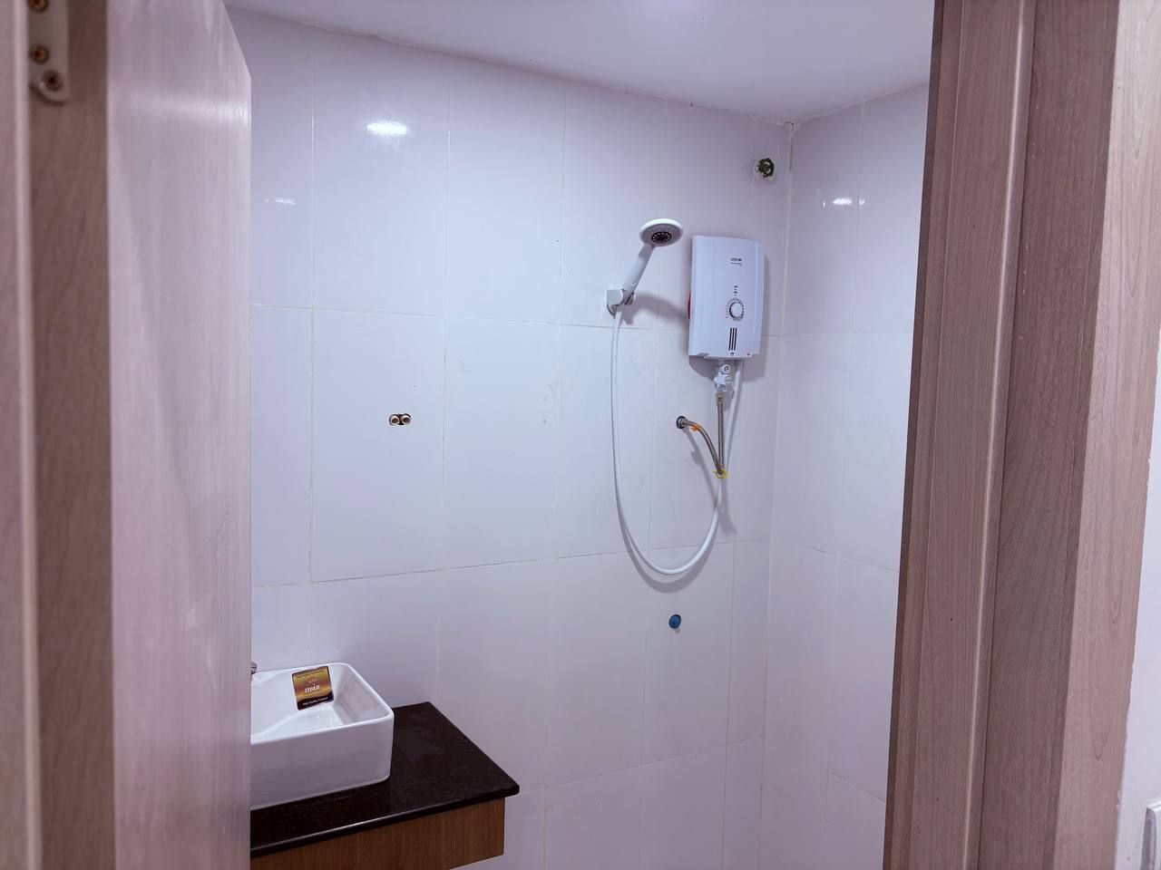 Pequeno espaço de chuveiro fechado, com parede revestida de azulejos brancos, chuveiro manual e painel de controle fixado na parede.