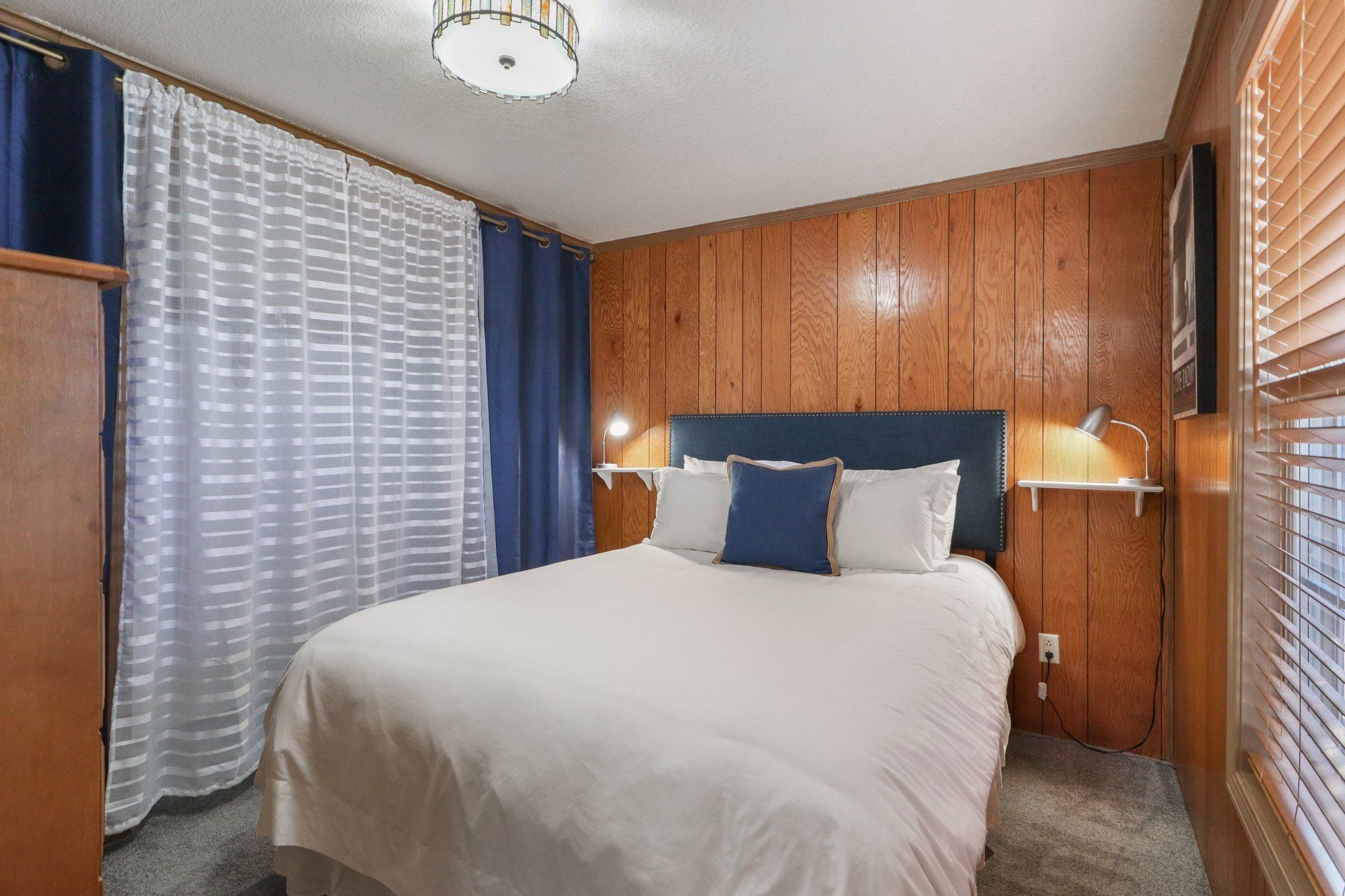 Ein Hotelzimmer mit einer Wandpaneelausstattung aus Holz, einem Bett mit weißer Bettwäsche, einem Nachttisch mit einer Lampe und einem Fenster mit Jalousien.