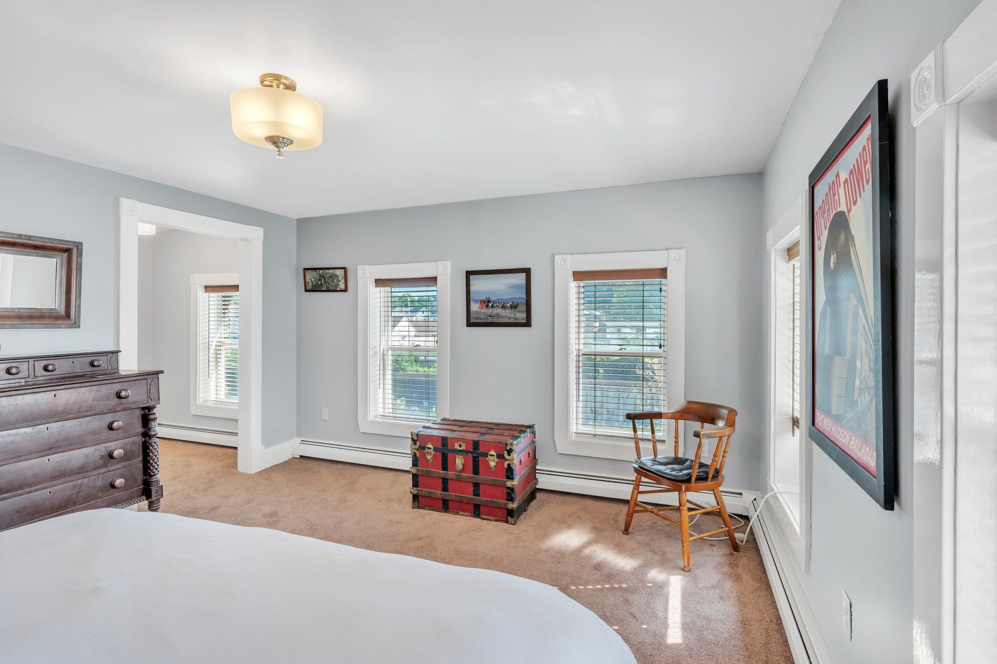 A bedroom with a bed, dresser, chest of drawers, a chest of drawers, a chair, a window with blinds, a picture frame, and a ceiling light fixture.