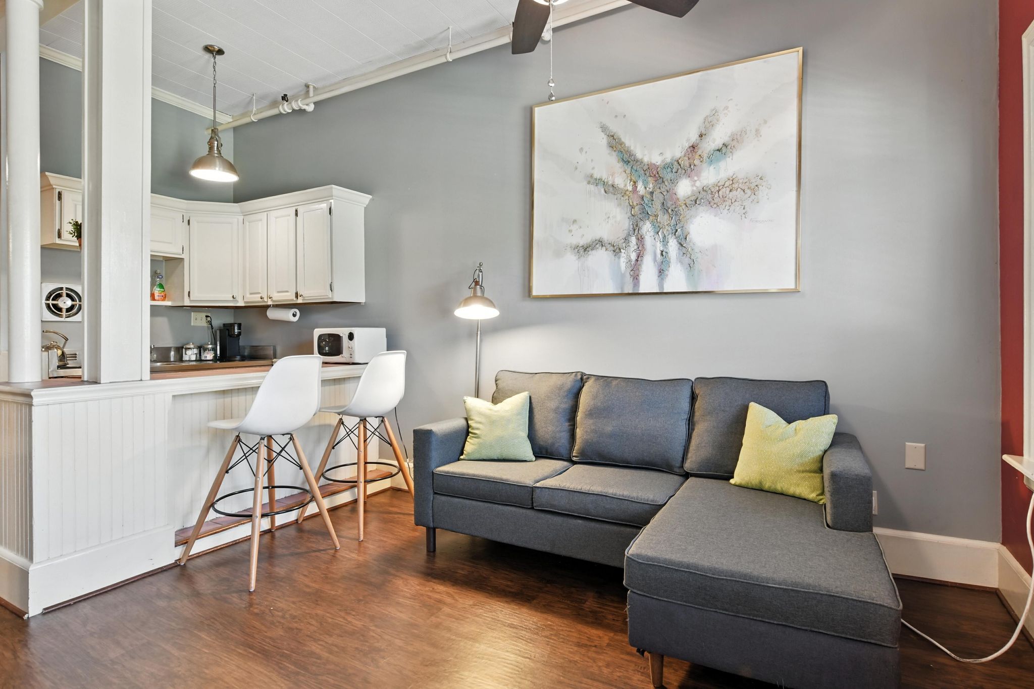 Un salon moderne avec un canapé d’angle gris, deux fauteuils blancs, un coin cuisine doté de meubles blancs et un ventilateur de plafond.