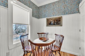A dining area with a white table and four wooden chairs, a window with a view of trees, and a wall with a patterned wallpaper. - Trainmaster's Inn - Viking house first floor (Palmer)