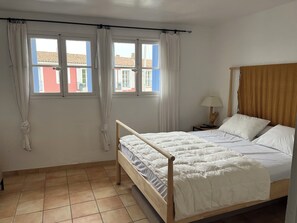 Et soveværelse med en træseng, hvide sengetøj, et natbord med en lampe og udsigt til bygninger gennem to vinduer.