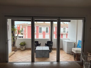 Ein Zimmer mit einer Schiebetür zum Außenbereich, wo eine Terrasse mit Tisch und Stühlen, ein Blumentopf und ein Stapel weißer Gegenstände zu sehen sind.