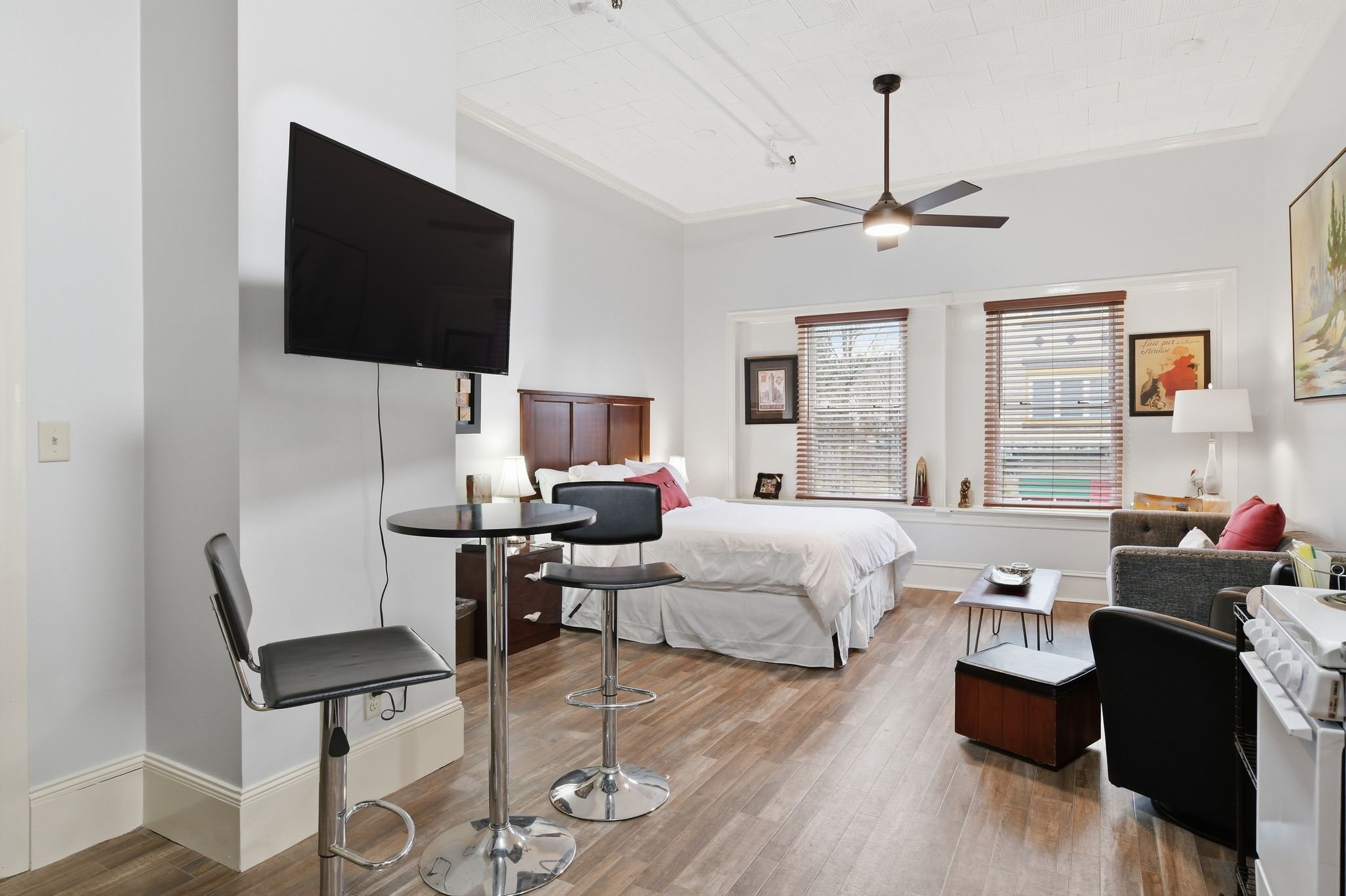 Een moderne hotelkamer met een flatscreen televisie, een plafondventilator, een bed met witte beddengoed, een bureau met een stoel en een raam met jaloezieën.