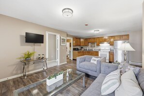 A modern living room with a flat-screen TV, a glass coffee table, and a grey sofa with decorative pillows. - Trainmaster's Inn - 1294 South Main, Apt 2 (Palmer)