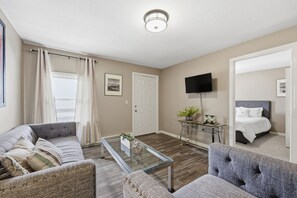 A well-lit, neutral-colored room with a sofa, armchair, and a glass coffee table. A flat-screen TV is mounted on the wall, and there's a door leading to another room. - Trainmaster's Inn - 1294 South Main, Apt 2 (Palmer)