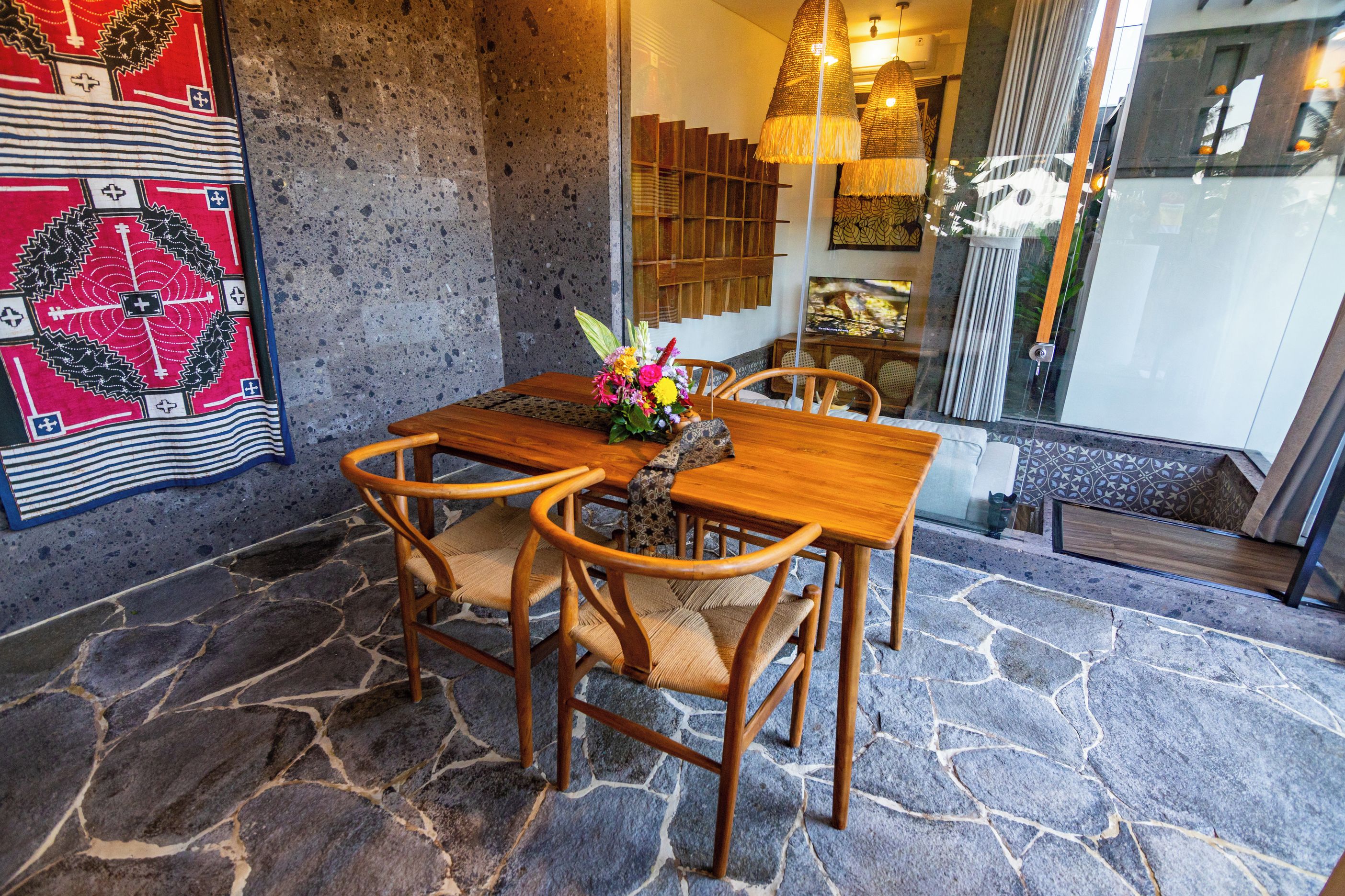 Uma sala de jantar com piso de pedra, mesa e cadeiras de madeira, um arranjo floral e um painel de parede estampado.
