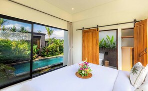 A bedroom with a large window, a bed with a floral arrangement, and a view of a swimming pool and greenery. - Sullaleca Villa (Gianyar)