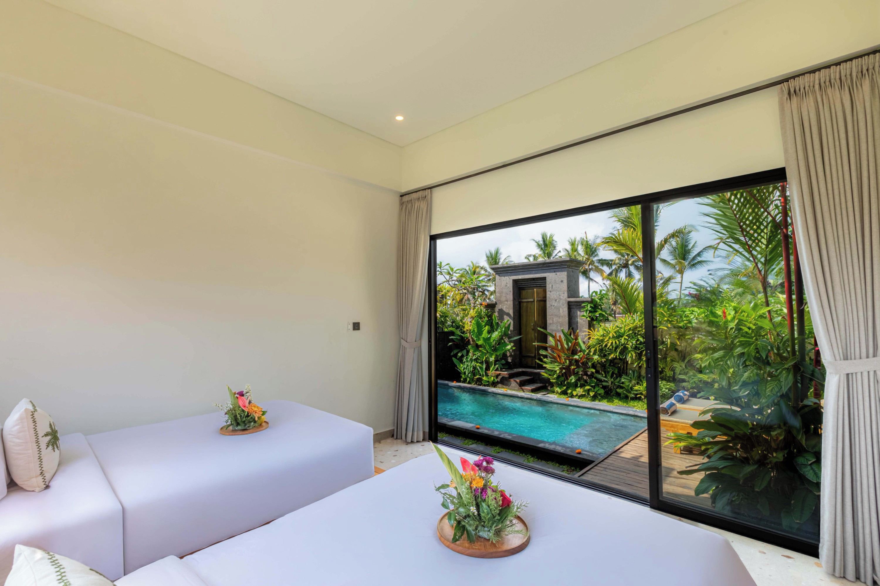 Camera d'albergo con due letti, un'ampia finestra che si affaccia su una piscina e piante tropicali, e un piccolo spazio esterno visibile attraverso la finestra.