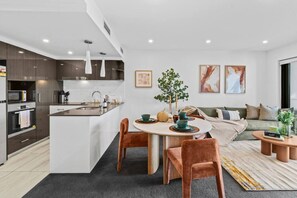 Cocina moderna con una isla central, comedor con mesa redonda y sillas, y sala de estar con sofá y cojines.
