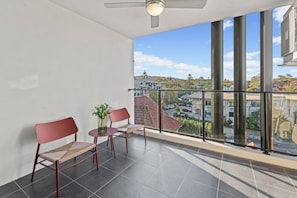 Un balcón con dos sillas y una mesita, grandes ventanales con vistas a una zona residencial.
