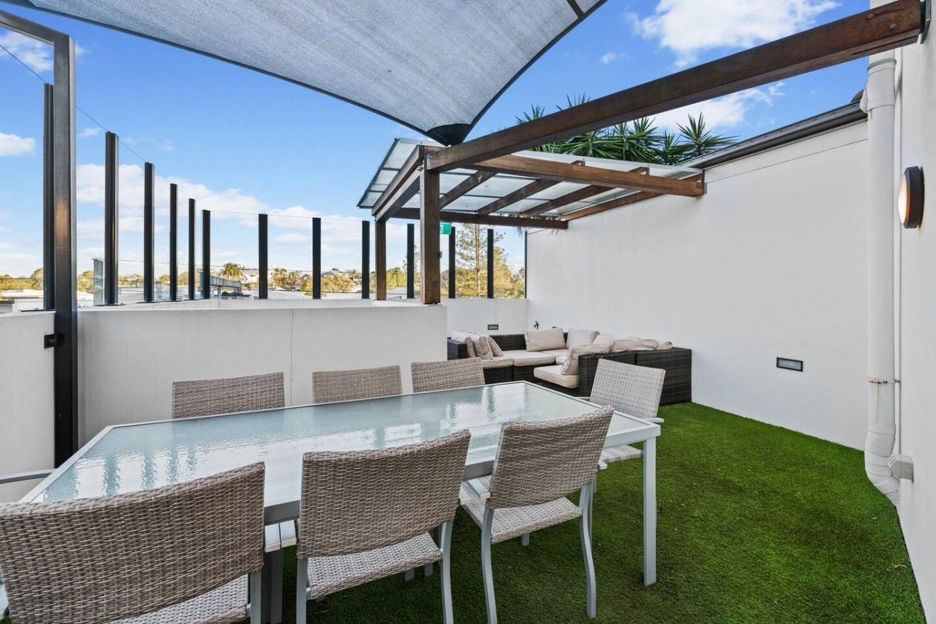 An outdoor patio with a glass-top table and wicker chairs, surrounded by a white wall and a green lawn.