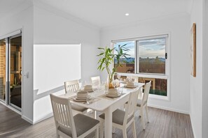 A dining area with a table set for four, white chairs, a potted plant, and a view of the outdoors through a large window. - Brand New4BR Home |Aircon Every Bedroom |GoodValue (White Rock)