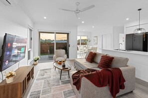 A modern living room with a flat-screen TV, a sofa, a coffee table, and a sliding glass door leading to a patio. - Brand New4BR Home |Aircon Every Bedroom |GoodValue (White Rock)
