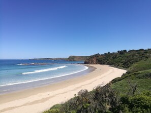 Uma praia costeira com céu claro, areal arenoso e paisagem montanhosa ao fundo.