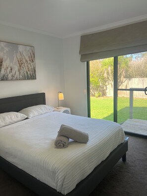 Quarto com uma cama grande, mesa de cabeceira, luminária e vista para um gramado através de uma porta de correr.
