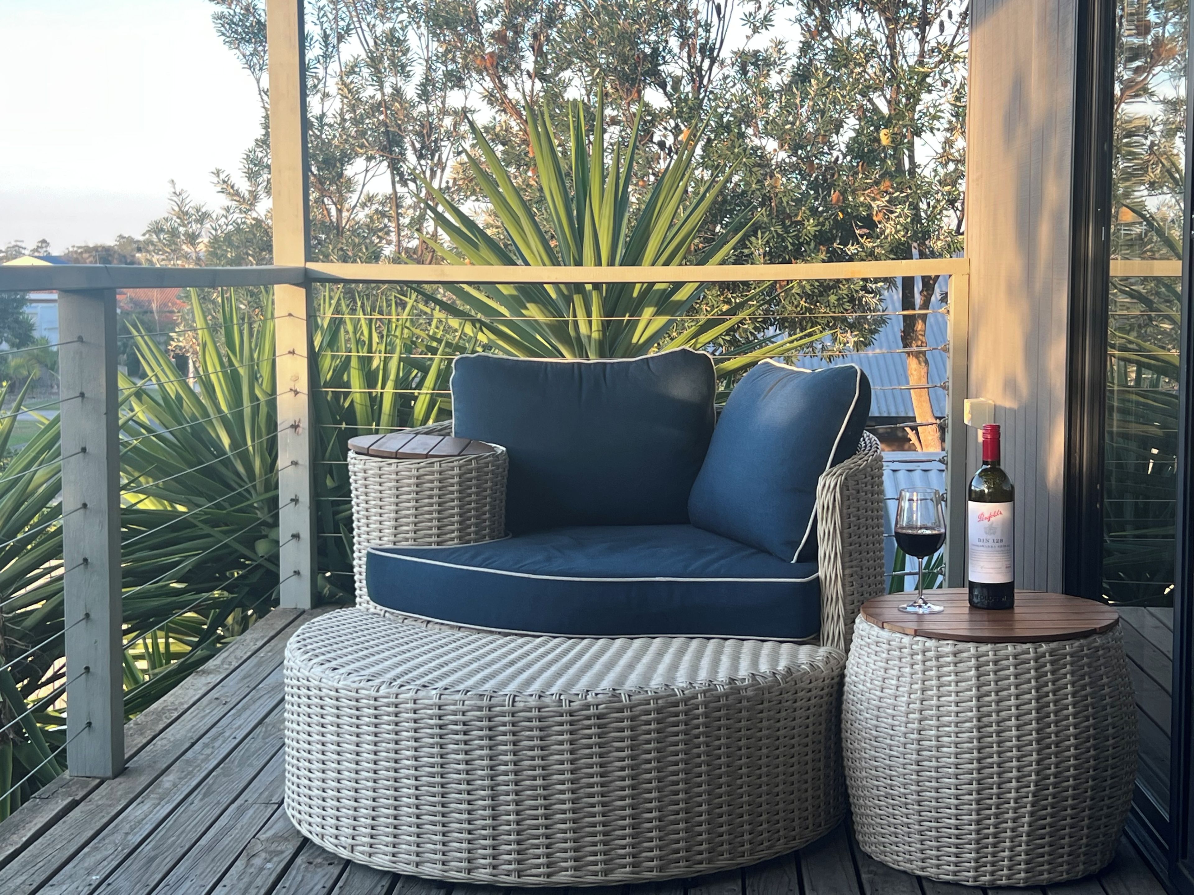 A wicker sofa with cushions, a round wicker table, and a bottle of wine on a wooden deck with a view of greenery.