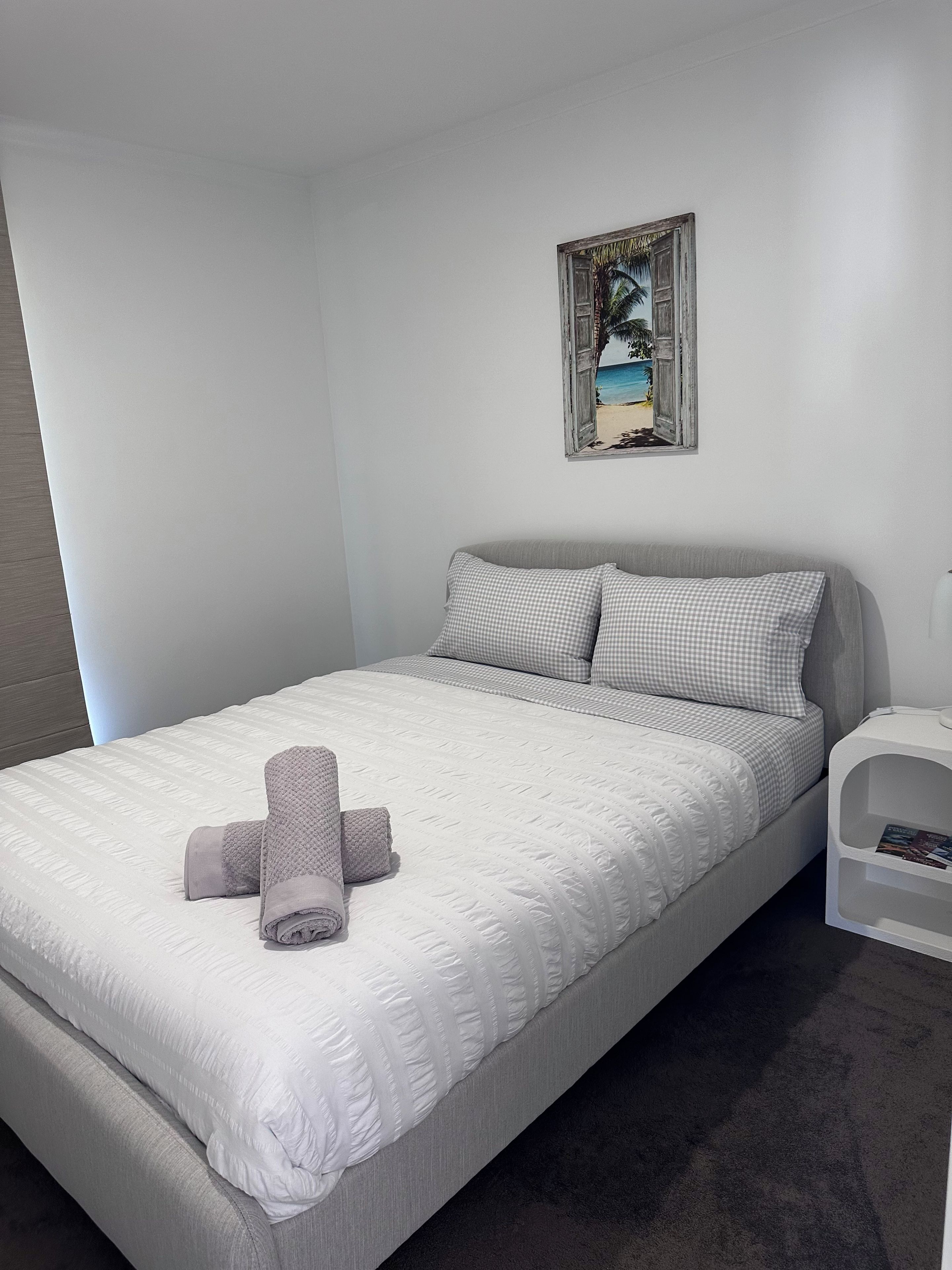 A neatly made bed with a checkered pillow and a rolled towel, a small bedside table with a magazine, and a wall-mounted painting of a beach scene.