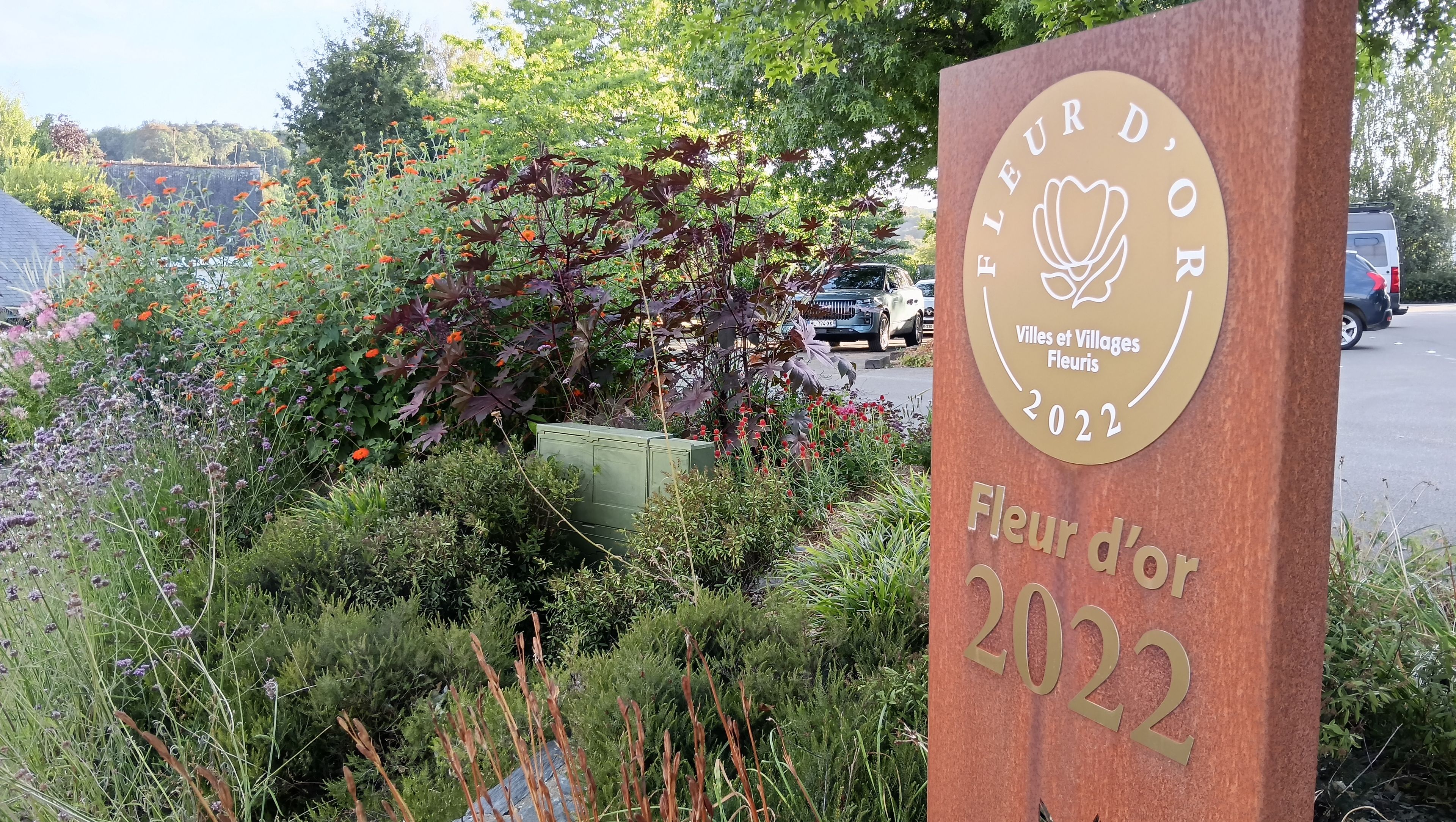 Une pancarte portant les inscriptions « Fleur d’Or 2022 » et « Villes et Villages Fleuris », entourée de verdure et de fleurs.