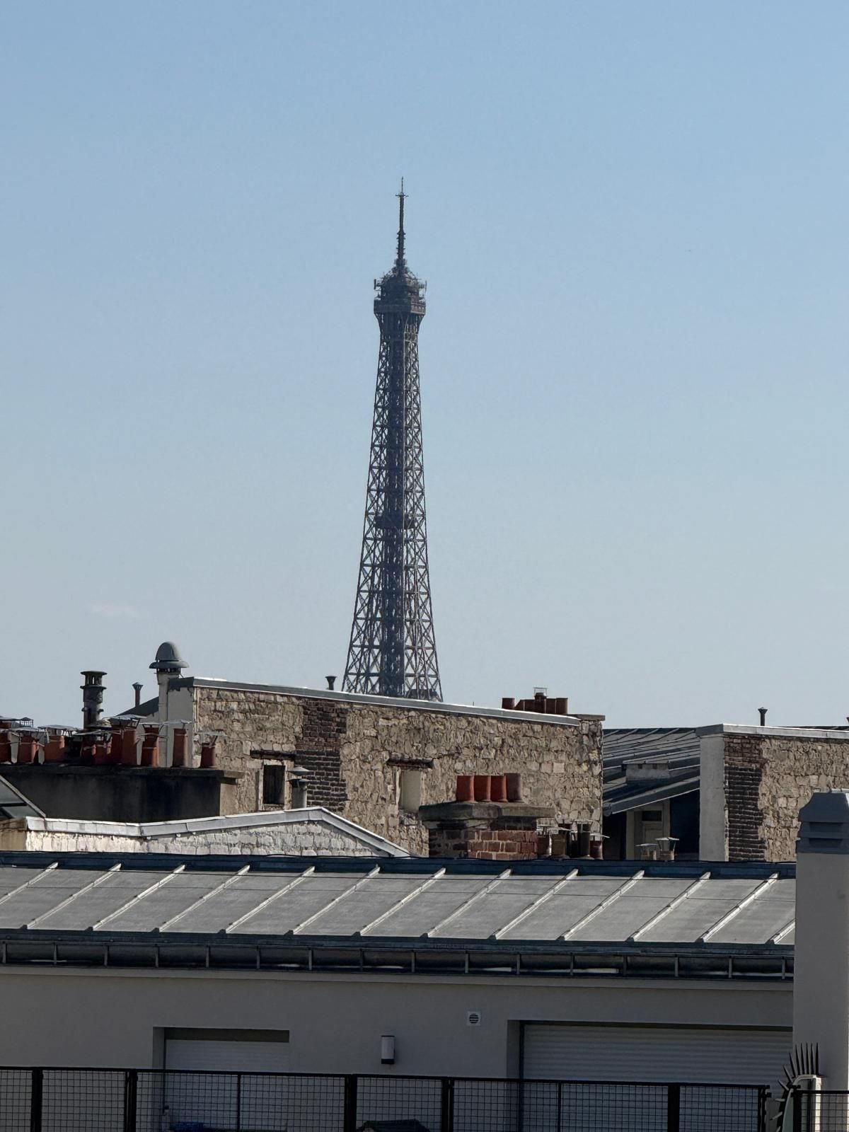 A tall metal tower with a cross at the top, surrounded by buildings with various rooftops and a clear sky.