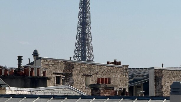 Uma torre de metal alta com uma cruz no topo, rodeada por edifícios com diferentes telhados e um céu limpo.