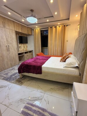Un dormitorio moderno con cama, televisión, armario y baño visible a través de una ventana.
