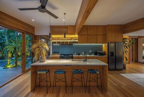 A modern kitchen with a central island, bar stools, wooden cabinetry, and a view of a tropical garden. - Bawa House (Pererenan)