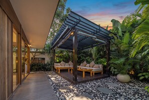 A covered outdoor seating area with a pergola, wooden deck, and surrounding tropical plants. - Bawa House (Pererenan)