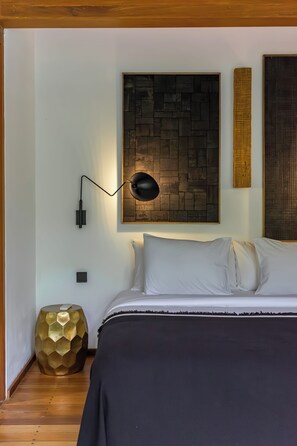 A bedroom with a bed, a wall-mounted lamp, a decorative wall piece, and a round gold-colored side table. - Bawa House (Pererenan)
