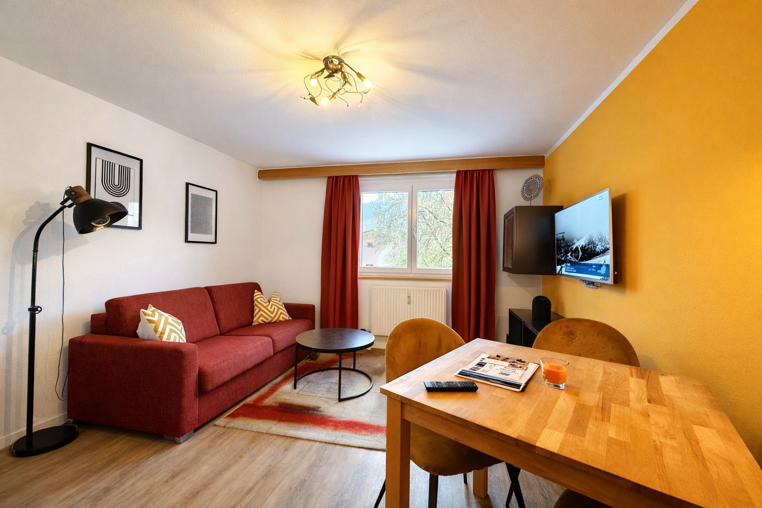 A furnished room with a red sofa, a wooden dining table, a TV mounted on the wall, and a ceiling fan.