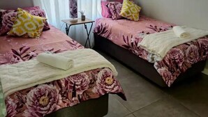 A room with two beds, each with floral bedding and pillows, a small table with a lamp, and a window with curtains. - TrueStar at TrueNorth (Pretoria)
