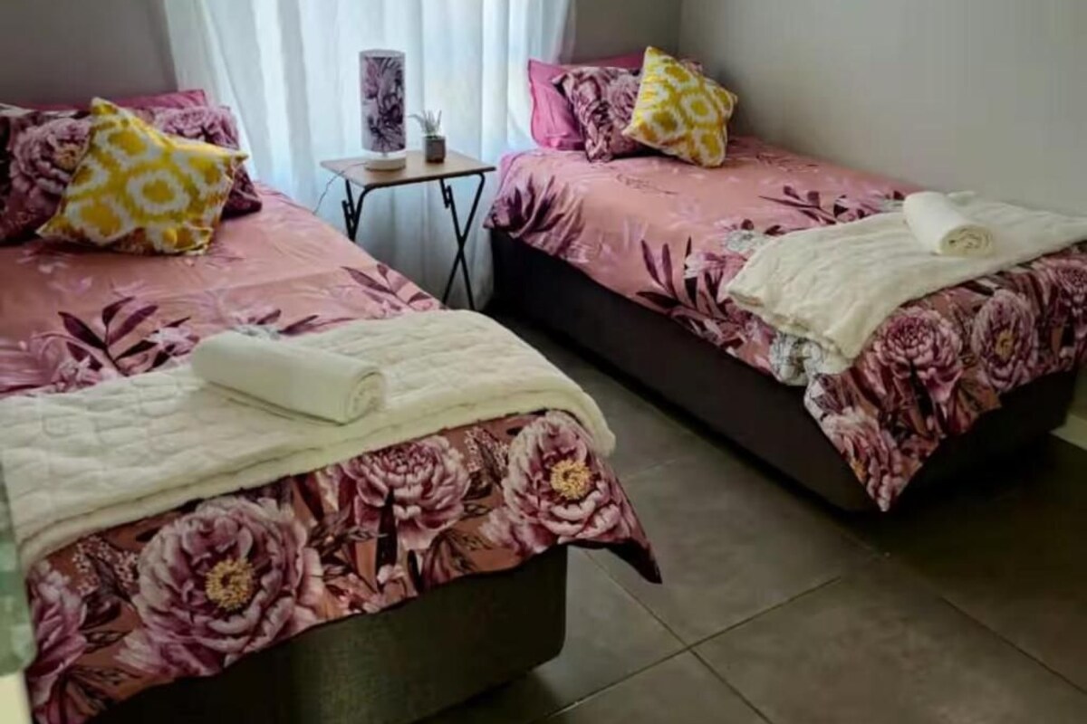 A room with two beds, each with floral bedding and pillows, a small table with a lamp, and a window with curtains.