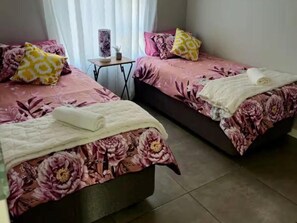 A room with two beds, each with floral bedding and pillows, a small table with a lamp, and a window with curtains. - TrueStar at TrueNorth (Pretoria)