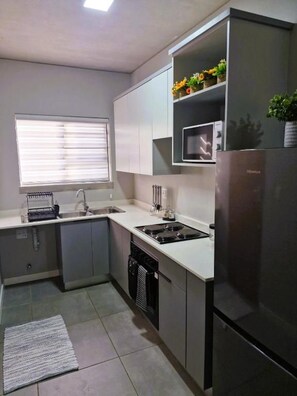 A compact kitchen with a built-in microwave, a stainless steel refrigerator, and a window with blinds. - TrueStar at TrueNorth (Pretoria)